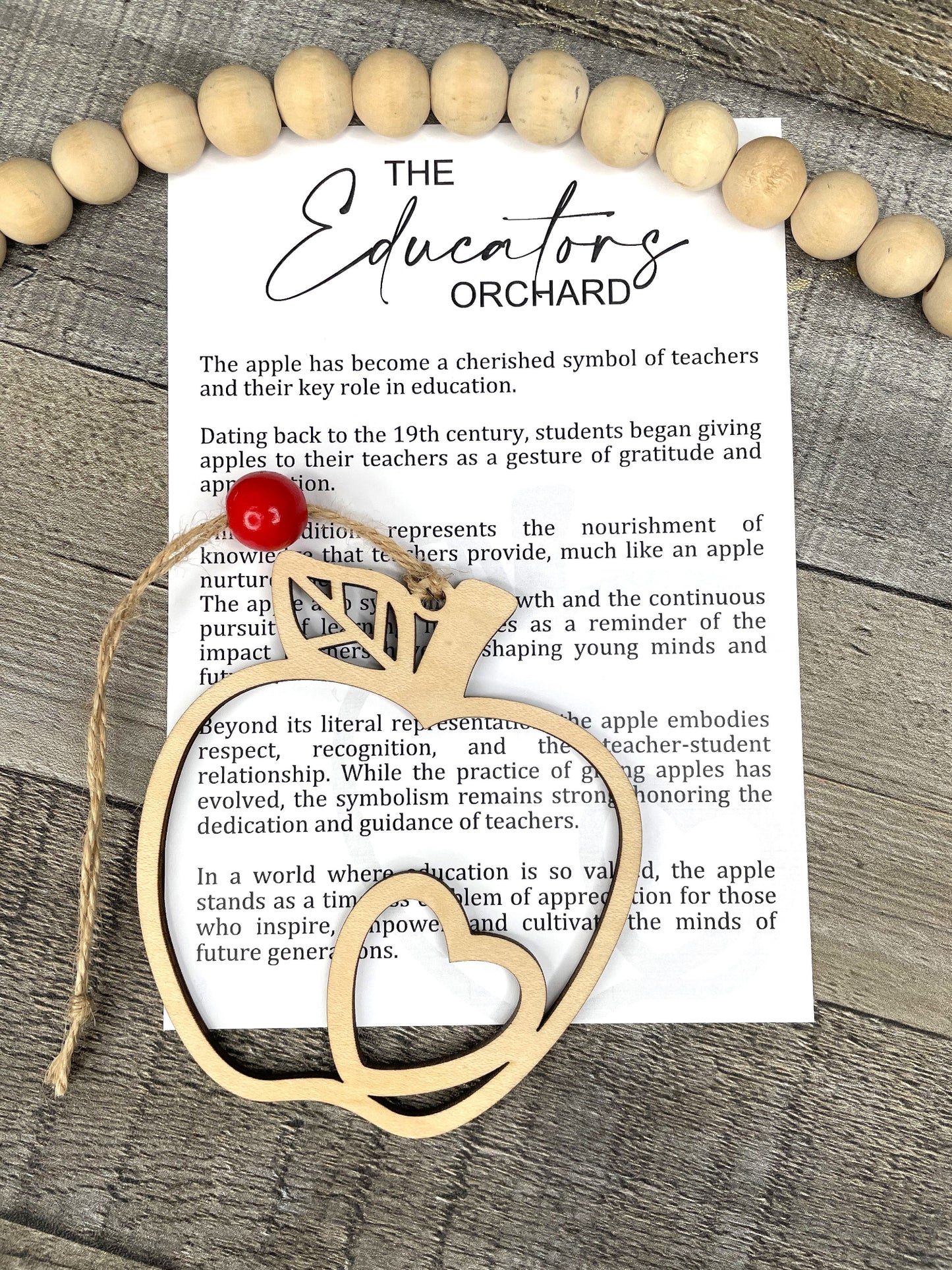 A story card charm with an apple symbol, attached to a natural wood bead, accompanied by a piece of paper with the text 'The Educators Orchard'.