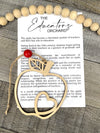 A story card charm with an apple symbol, attached to a natural wood bead, accompanied by a piece of paper with the text 'The Educators Orchard'.
