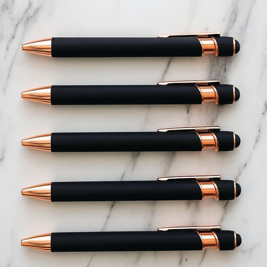 Five black and rose gold pens arranged on a marble surface