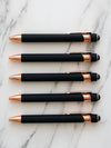 Five black and rose gold pens arranged on a marble surface