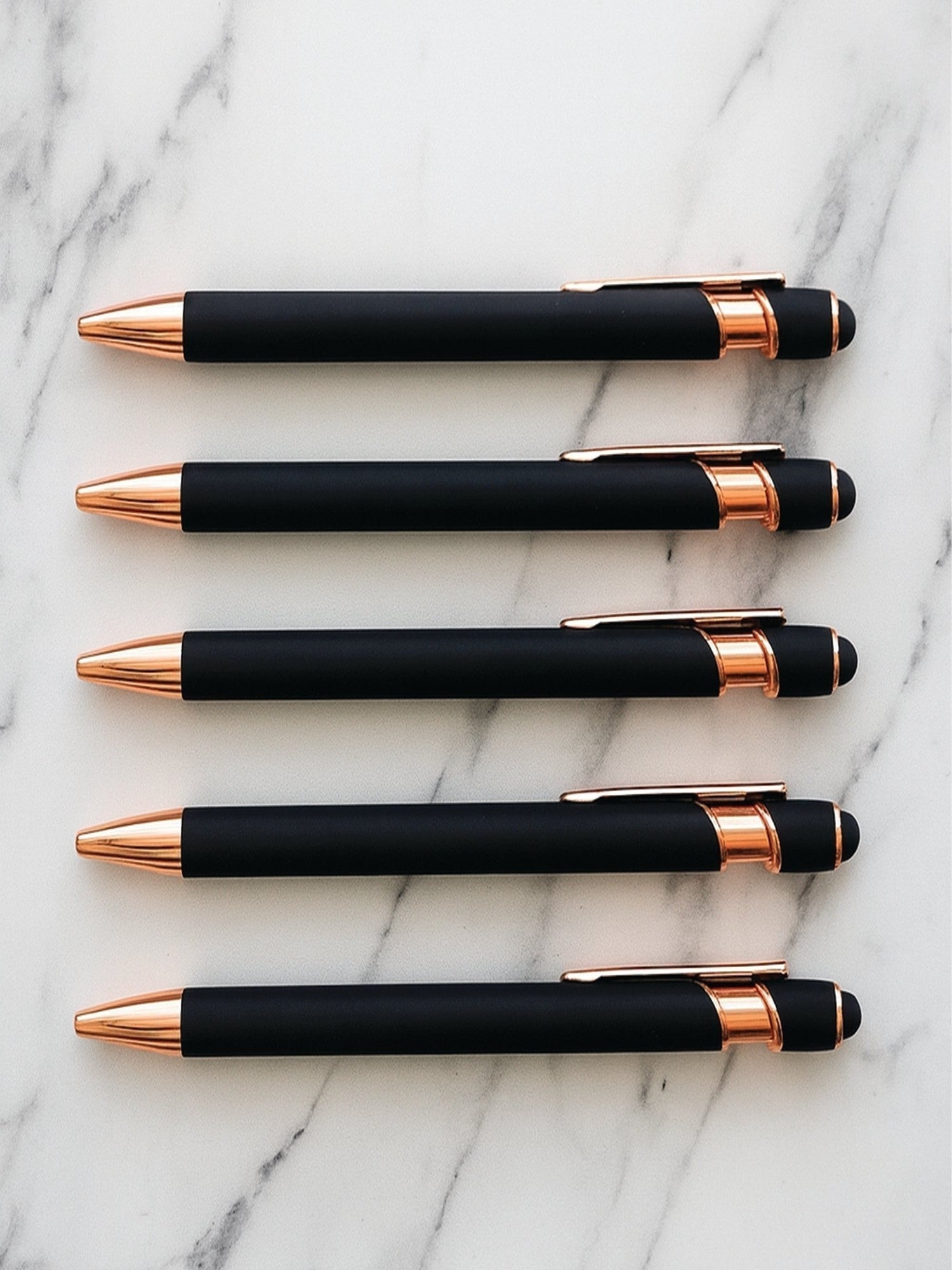 Five black and rose gold pens arranged on a marble surface