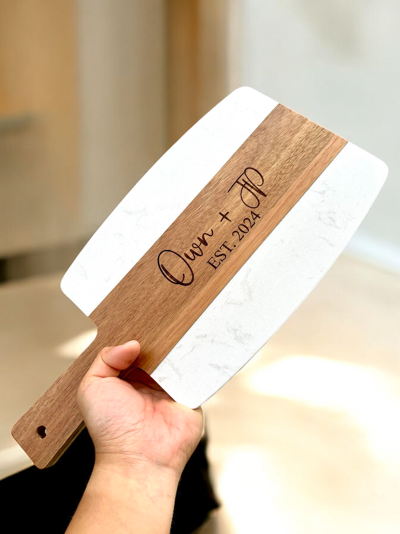 A hand holding a serving board with a white marble top and a laser engraved name 'Quinn' on the acacia wood part.