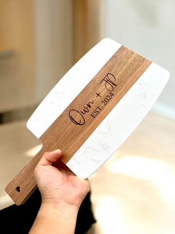 A hand holding a serving board with a white marble top and a laser engraved name 'Quinn' on the acacia wood part.