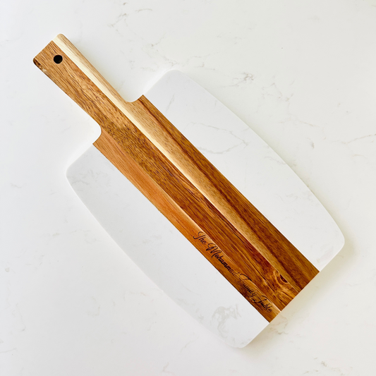 A personalized cheese board made of wood and marble, with a white marble surface and a wooden handle, resting on a marble surface.